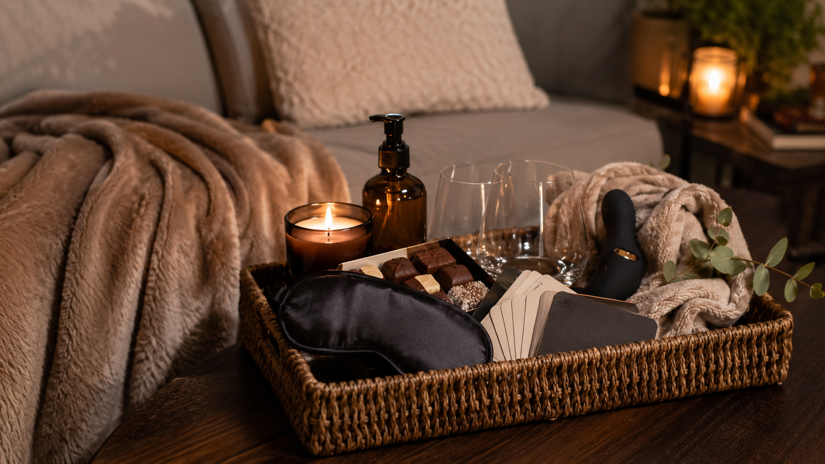 At-home adult date night gift basket on a coffee table with a candle, massage oil, glasses, chocolates, cards, and a soft blanket.