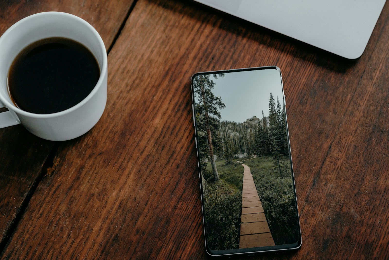 A real smartphone on a wooden desk showing a nature wallpaper beside a coffee cup and laptop.